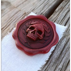 May include: A circular dark red wax seal featuring a raised relief of a human skull centered within a laurel wreath. The wax seal has an irregular, scalloped edge and a smooth, glossy finish, displaying intricate details of the skull and leaves.