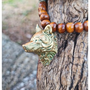 Op de afbeelding: Een armband met gladde, ronde, gepolijste bruine houten kralen, geaccentueerd door een gedetailleerde, driedimensionale metalen wolfskopbedel met een verweerde, antieke messing afwerking en subtiele groene patina-accenten.
