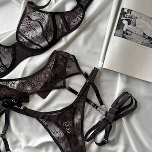 Könnte beinhalten: Ein Set aus dunkelbrauner Spitzenwäsche, bestehend aus BH, String-Tanga und Harness. Die Wäsche ist auf einem weißen, seidigen Stoff drapiert. Eine Zeitschrift mit Schwarz-Weiß-Foto ist teilweise sichtbar.