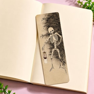 Puede incluir: Un marcapáginas rectangular con una ilustración en tinta negra y blanca de un esqueleto de pie en un bosque de bambú. El esqueleto sostiene una pequeña linterna en una mano. El marcapáginas tiene un fondo beige claro y esquinas redondeadas.