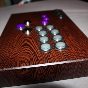 Puede incluir: Un mando de juego arcade de madera con un acabado de vetas de madera marrón oscuro. Cuenta con varios botones en varios colores, incluyendo morado y gris, dispuestos para el juego. El mando es rectangular y parece nuevo.