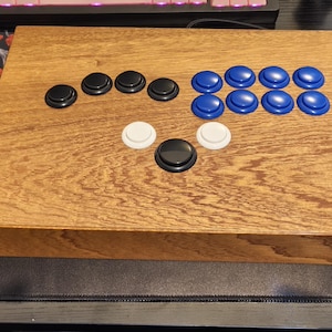 Puede incluir: Un mando de lucha arcade de madera con un acabado marrón claro. Cuenta con botones negros, blancos y azules dispuestos en un diseño estándar. El mando de lucha es rectangular y se encuentra sobre una superficie negra.