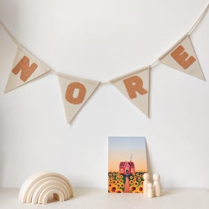 May include: A cream-coloured banner with the word "NORE" in brown letters. Beneath, a wooden rainbow stacker, a painting of a red house in a sunflower field, and wooden peg dolls are displayed.