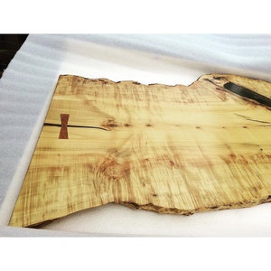 May include: A large, natural wood slab table top with a live edge. The wood is a light yellow color with dark brown streaks. A black epoxy resin inlay runs down the center of the table top.