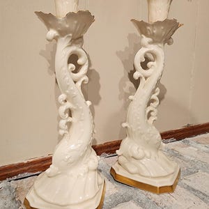 May include: Two white ceramic dolphin candlesticks with gold edging. The candlesticks have an ornate design and are suitable for standard candles. A decorative home decor item.