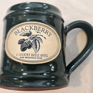 May include: A dark green ceramic mug with a glossy finish and a curved handle. An oval tan label on the front features a black illustration of a blackberry branch and the text: BLACKBERRY A COUNTRY HOUSE HOTEL AND MOUNTAIN CLUB.