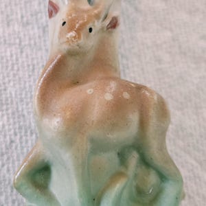 May include: A ceramic figurine of a deer standing in stylized green grass. The deer is light brown with white spots on its back and dark-tipped antlers. The base is a pale green, and the upper portion of the piece features a smooth, off-white finish.