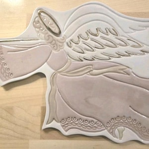 May include: A ceramic wall plaque shaped like an angel playing a trumpet. The angel features a white background with soft pink accents on the robe and wings, highlighted by metallic gold trim on the halo, wings, trumpet, and hem details.