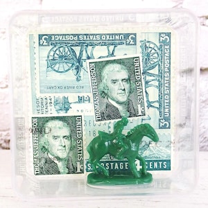 Könnte beinhalten: Ein durchsichtiger Kunststoffbehälter mit alten US-amerikanischen Briefmarken mit Thomas Jefferson und einem grünen Spielzeugsoldaten auf einem Pferd. Die Briefmarken haben einen Wert von 3 Cent, mit dem Text "Red River Ox Cart".