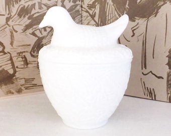 May include: A white milk glass lidded jar featuring a bird-shaped finial on the lid. The body of the jar has a textured, embossed pattern across its surface. The lid sits securely on top of the rounded container, creating a cohesive piece.