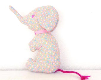 May include: A stuffed elephant toy made of tan fabric with a small blue, pink, and white floral pattern. The elephant features a bright pink ribbon collar, a pink stitched eye, and a long, thin, braided bright pink tail with a tassel end.