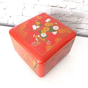 May include: A red, square box with a glossy finish. The lid is decorated with a floral design featuring white, yellow, and orange flowers. Gold geometric patterns frame the floral arrangement. The box is likely used for storage or display.