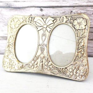 May include: A vintage silver-toned double photo frame with oval openings. The frame is decorated with floral and bow motifs. The frame is made of metal and has a slightly curved shape.