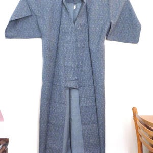 May include: A blue kimono featuring a subtle, speckled pattern throughout the fabric. The garment has wide, straight sleeves, a V-neckline, and a long, flowing silhouette that hangs straight down from the shoulders, displayed on a wooden hanger.