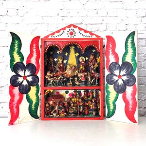 Könnte beinhalten: Eine rot handgeschnitzte Krippe mit zwei Ebenen von Figuren. Die Szene wird von weißen Tafeln mit roten, grünen und schwarzen floralen Designs umrahmt. Der Hintergrund ist eine weiße Backsteinmauer.