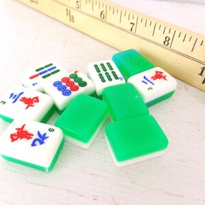 Puede incluir: Un conjunto de fichas de Mahjong con caras blancas y bordes verdes, que muestran varios s&iacute;mbolos en rojo, azul y verde. Una regla de madera es parcialmente visible en el fondo, lo que sugiere un juego o un contexto artesanal.