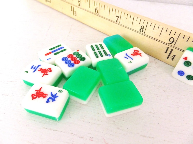 Puede incluir: Un conjunto de fichas de Mahjong con caras blancas y bordes verdes, que muestran varios s&iacute;mbolos en rojo, azul y verde. Una regla de madera es parcialmente visible en el fondo, lo que sugiere un juego o un contexto artesanal.