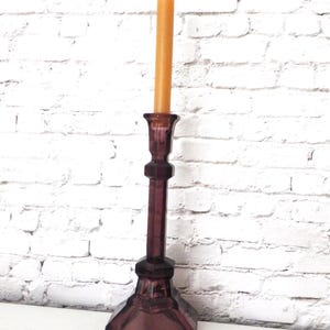 May include: A tall, antique-style candle holder in a deep purple hue. The candlestick features a hexagonal base, a slender stem, and a cup-shaped top holding a single, unlit, orange candle. The background is a white brick wall.