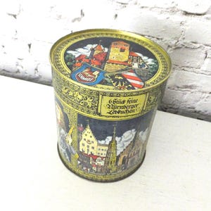 May include: Vintage round tin with a gold rim and a detailed illustration of a European town. The tin features a castle, buildings, and text in German. The tin is likely for biscuits or other treats.