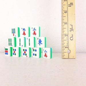 Puede incluir: Un conjunto de fichas de Mahjong blancas con bordes verdes y varios s&iacute;mbolos y caracteres en rojo, azul y verde. Una regla de madera con medidas en el fondo, mide aproximadamente 13 cm.