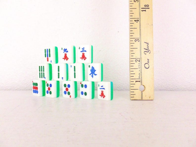 Puede incluir: Un conjunto de fichas de Mahjong blancas con bordes verdes y varios s&iacute;mbolos y caracteres en rojo, azul y verde. Una regla de madera con medidas en el fondo, mide aproximadamente 13 cm.