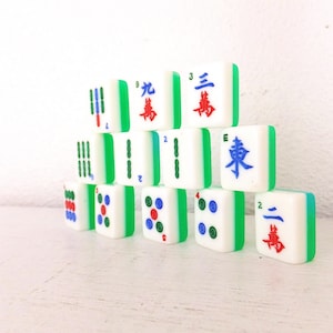 Puede incluir: Un conjunto de fichas de Mahjong blancas con bordes verdes, apiladas en forma de pir&aacute;mide. Las fichas presentan s&iacute;mbolos y n&uacute;meros rojos, azules y verdes. Las piezas del juego est&aacute;n dispuestas sobre una superficie blanca.
