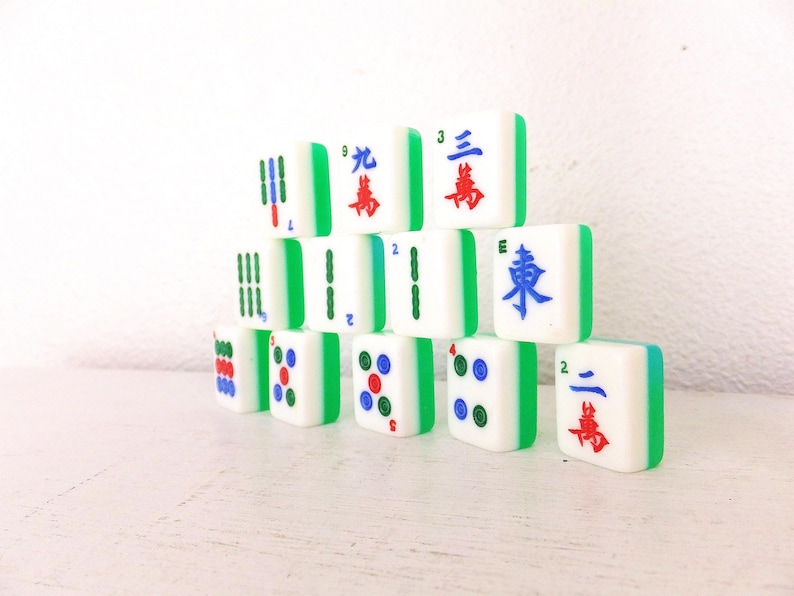 Puede incluir: Un conjunto de fichas de Mahjong blancas con bordes verdes, apiladas en forma de pir&aacute;mide. Las fichas presentan s&iacute;mbolos y n&uacute;meros rojos, azules y verdes. Las piezas del juego est&aacute;n dispuestas sobre una superficie blanca.