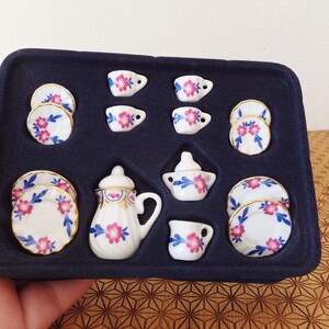 May include: A miniature tea set in a black velvet box. The set includes a teapot, creamer, sugar bowl, four teacups, and four saucers. The porcelain is white with blue and pink floral designs.