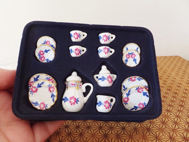 May include: A miniature tea set in a black velvet box. The set includes a teapot, creamer, sugar bowl, four teacups, and four saucers. The porcelain is white with blue and pink floral designs.