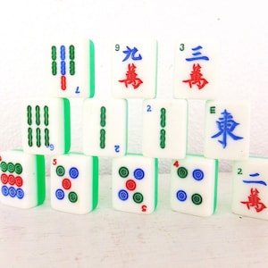 Puede incluir: Un conjunto de fichas de Mahjong blancas con bordes verdes. Las fichas muestran varios s&iacute;mbolos, n&uacute;meros y caracteres chinos en rojo, azul y verde. Las fichas est&aacute;n apiladas, mostrando los componentes del juego.