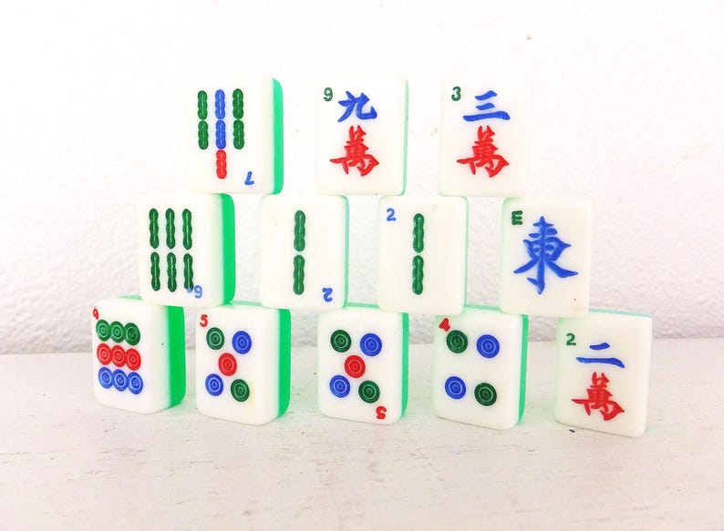 Puede incluir: Un conjunto de fichas de Mahjong blancas con bordes verdes. Las fichas muestran varios s&iacute;mbolos, n&uacute;meros y caracteres chinos en rojo, azul y verde. Las fichas est&aacute;n apiladas, mostrando los componentes del juego.
