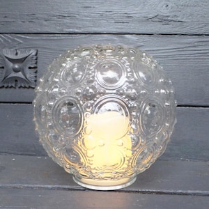 May include: A clear glass globe-shaped candle holder with a textured pattern of circles and ovals. The candle holder sits on a white base and contains a lit, yellow candle. The background is a dark gray wooden surface.