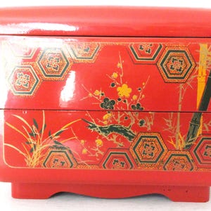 May include: A red, two-tiered Japanese bento box with a glossy finish. The box features floral and bamboo designs with gold and black accents. The design includes hexagonal patterns and botanical illustrations.