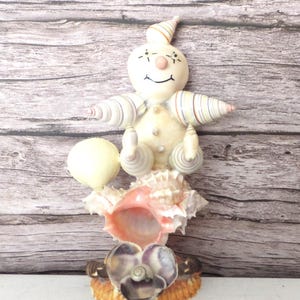 May include: A whimsical seashell sculpture featuring a smiling, cream-colored figure with striped arms and a pointed hat. The figure sits atop a large, pink-hued seashell, with a smaller shell base. A yellow sphere is held in the figure's hand.
