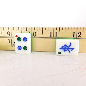 Puede incluir: Primer plano de tres fichas de Mahjong. Dos fichas son blancas con bordes verdes y s&iacute;mbolos azules y verdes. La tercera ficha es blanca con bordes verdes y un s&iacute;mbolo rojo. Una regla de madera est&aacute; en el fondo.