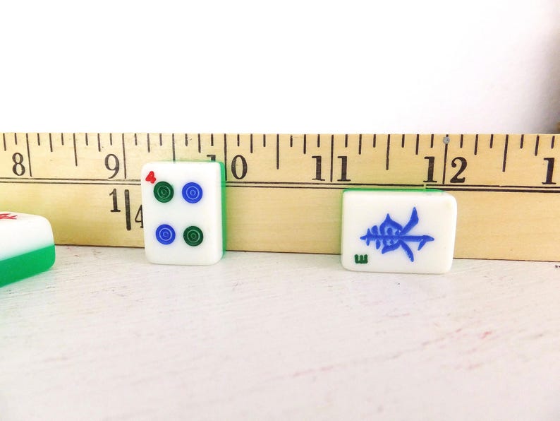Puede incluir: Primer plano de tres fichas de Mahjong. Dos fichas son blancas con bordes verdes y s&iacute;mbolos azules y verdes. La tercera ficha es blanca con bordes verdes y un s&iacute;mbolo rojo. Una regla de madera est&aacute; en el fondo.