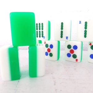 Puede incluir: Una colecci&oacute;n de fichas de Mahjong blancas y verdes. Algunas fichas tienen dorso verde, mientras que otras muestran varios s&iacute;mbolos y n&uacute;meros en verde, rojo y azul. Las fichas est&aacute;n apiladas y dispuestas sobre una superficie blanca.