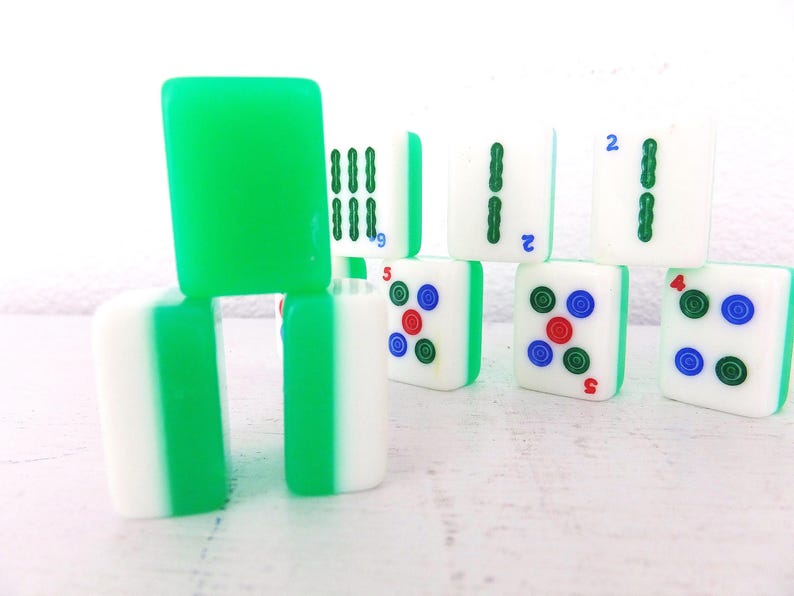 Puede incluir: Una colecci&oacute;n de fichas de Mahjong blancas y verdes. Algunas fichas tienen dorso verde, mientras que otras muestran varios s&iacute;mbolos y n&uacute;meros en verde, rojo y azul. Las fichas est&aacute;n apiladas y dispuestas sobre una superficie blanca.