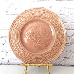 May include: A round, peach-colored ceramic plate with a raised relief design of a classical figure. The plate has a textured border and is displayed on a gold-colored stand. The plate is a decorative item.