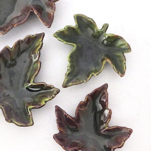 Peut inclure: Quatre petits plats en céramique en forme de feuilles, dans des tons de vert et de marron. Les plats ont une finition brillante et sont disposés sur une surface blanche. Les plats en forme de feuilles sont un élément décoratif.
