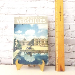Puede incluir: Un libro de viaje vintage titulado "The Sights of Versailles" con una imagen en color del palacio y el estanque reflectante. El libro está apoyado en un soporte dorado junto a una regla de madera con medidas en centímetros.