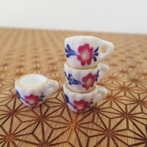 May include: A stack of four miniature teacups with floral designs in shades of pink, blue, and white. The cups are stacked on a wooden surface with a geometric pattern.