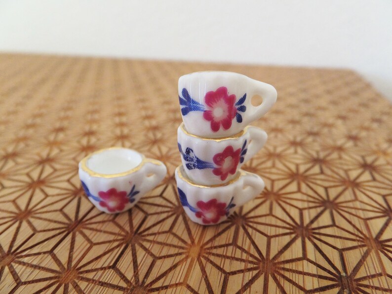 May include: A stack of four miniature teacups with floral designs in shades of pink, blue, and white. The cups are stacked on a wooden surface with a geometric pattern.