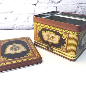 May include: A rectangular brown metal storage box with a matching lid. The box and lid feature ornate gold and black designs. The box is open, revealing a silver interior. The lid is placed next to the box.