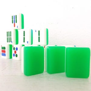 Puede incluir: Un conjunto de fichas de Mahjong, verdes y blancas. Las fichas muestran s&iacute;mbolos y n&uacute;meros en rojo, azul y verde. Algunas fichas est&aacute;n apiladas en forma de pir&aacute;mide, otras est&aacute;n dispuestas en fila.