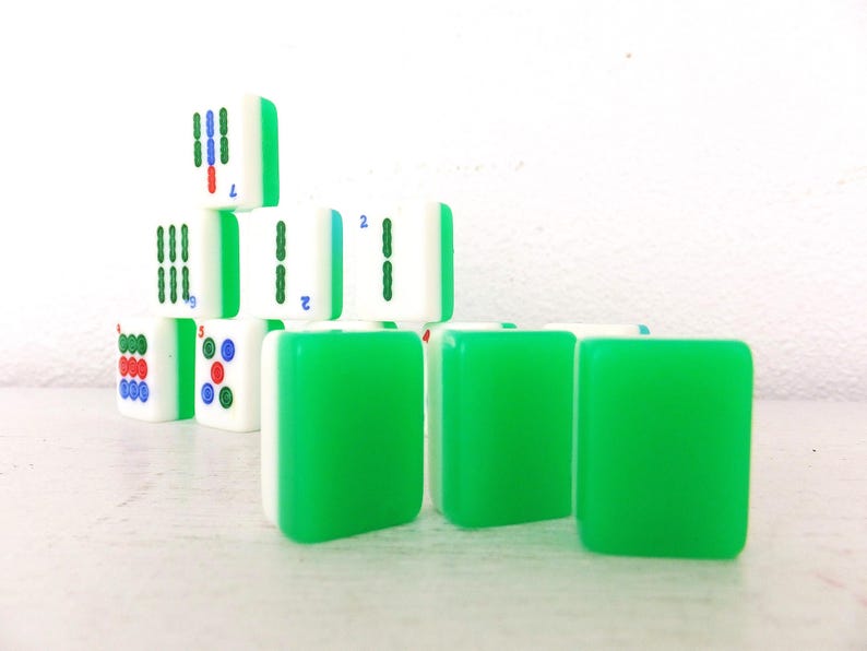 Puede incluir: Un conjunto de fichas de Mahjong, verdes y blancas. Las fichas muestran s&iacute;mbolos y n&uacute;meros en rojo, azul y verde. Algunas fichas est&aacute;n apiladas en forma de pir&aacute;mide, otras est&aacute;n dispuestas en fila.