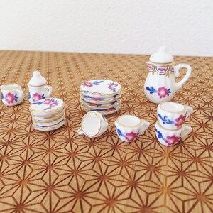 May include: A miniature tea set featuring a white teapot, cups, and saucers decorated with a floral design in shades of pink and blue. The set is displayed on a wooden surface with a geometric pattern.