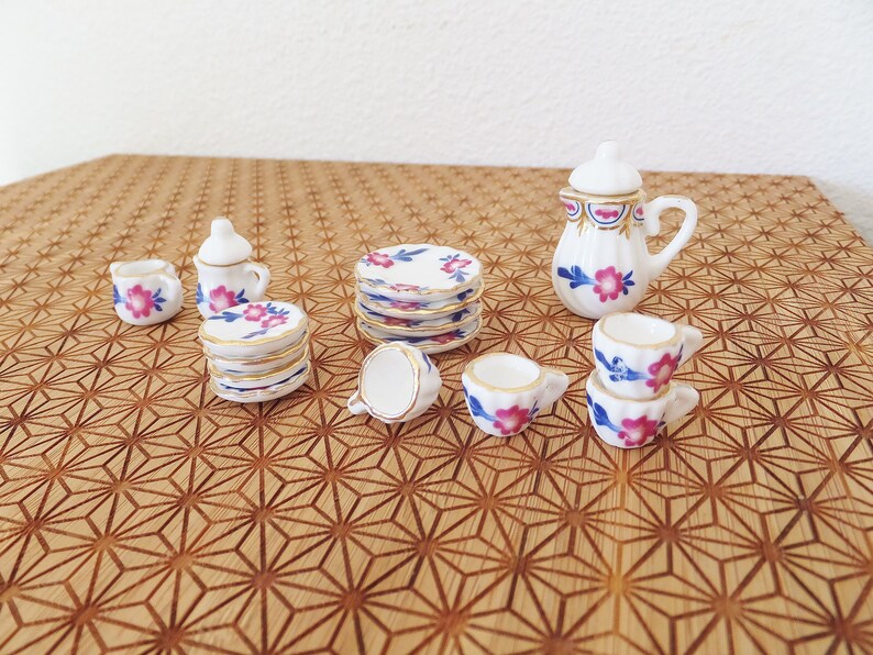 May include: A miniature tea set featuring a white teapot, cups, and saucers decorated with a floral design in shades of pink and blue. The set is displayed on a wooden surface with a geometric pattern.