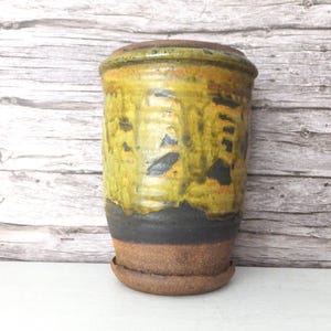 May include: A handmade ceramic plant pot with a lid. The pot has a textured surface with yellow, black, and brown tones. The cylindrical pot sits on a matching saucer, ideal for indoor plants.