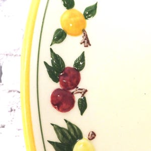 May include: A cream-colored ceramic plate featuring a hand-painted border of colorful fruit including a yellow lemon, red cherries, and green leaves. The plate has a bright yellow rim and a thin green line accenting the inner edge of the border.